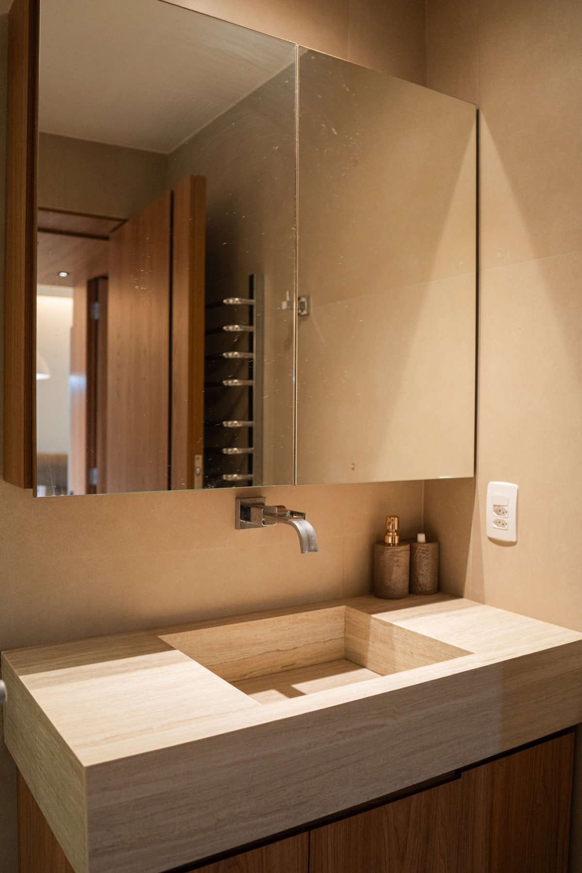 A modern bathroom features a sleek stone sink integrated into a wooden vanity, offering a minimalist design. A large mirror is mounted above the sink, reflecting the warm tones of the surrounding walls. The faucet is stylishly positioned, enhancing the contemporary look.