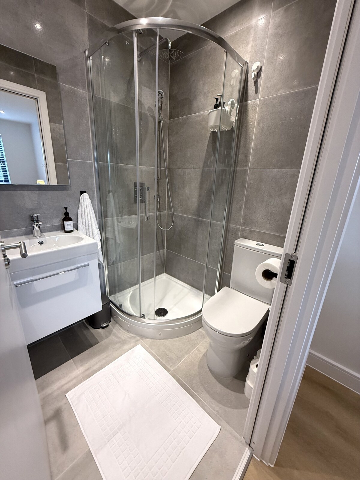 A modern bathroom features sleek gray tiles and glass shower enclosure. A white vanity with a sink and storage is positioned beside a toilet. A plush white bath mat is placed on the floor, enhancing the clean aesthetic. Natural light enters through a window.