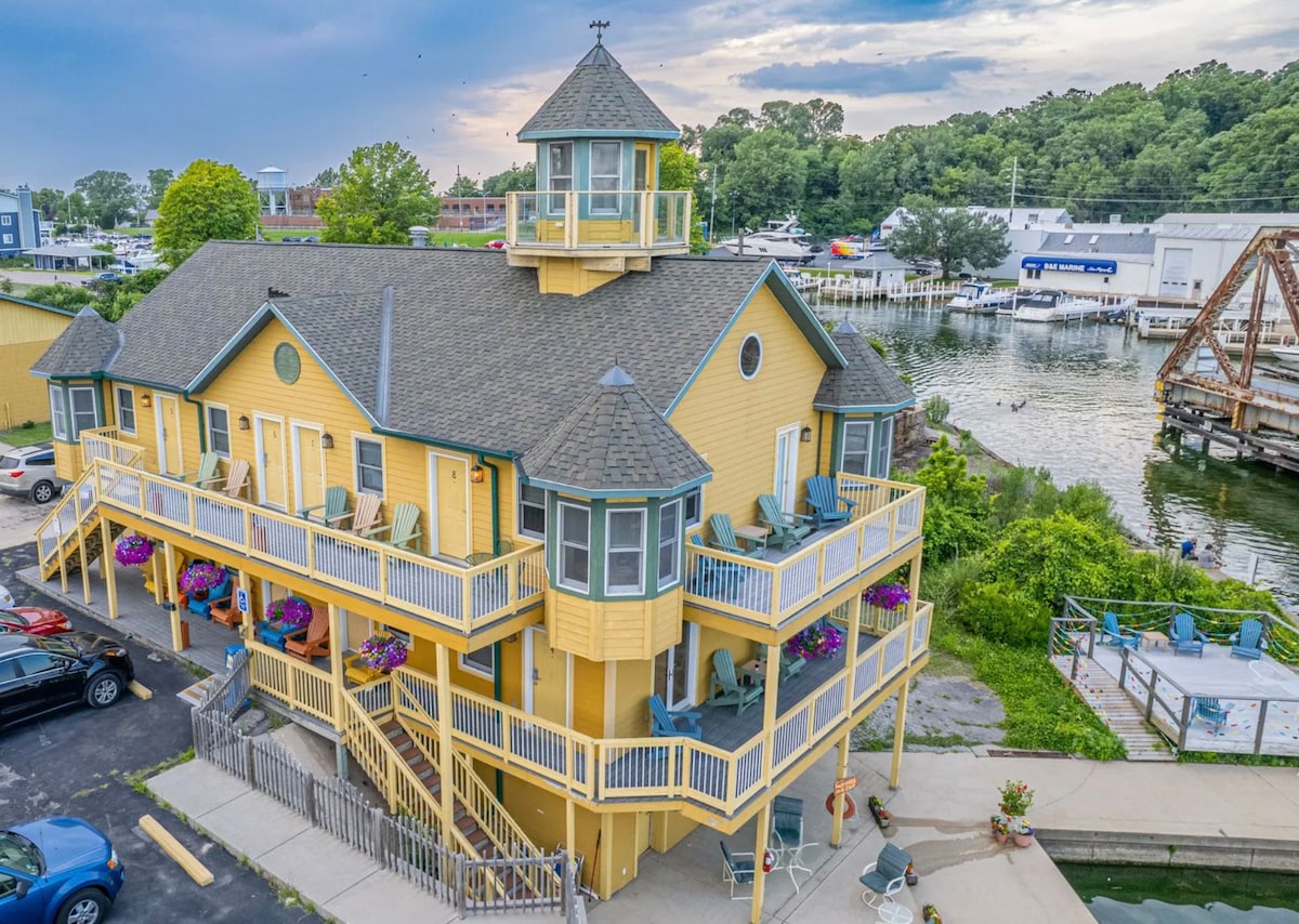 A multi-level yellow building with a distinctive cupola is positioned near a marina. Balconies adorned with colorful flower planters provide seating, enhancing the outdoor space. The surrounding area features parked vehicles and a waterfront view, contributing to the overall charm of the location.