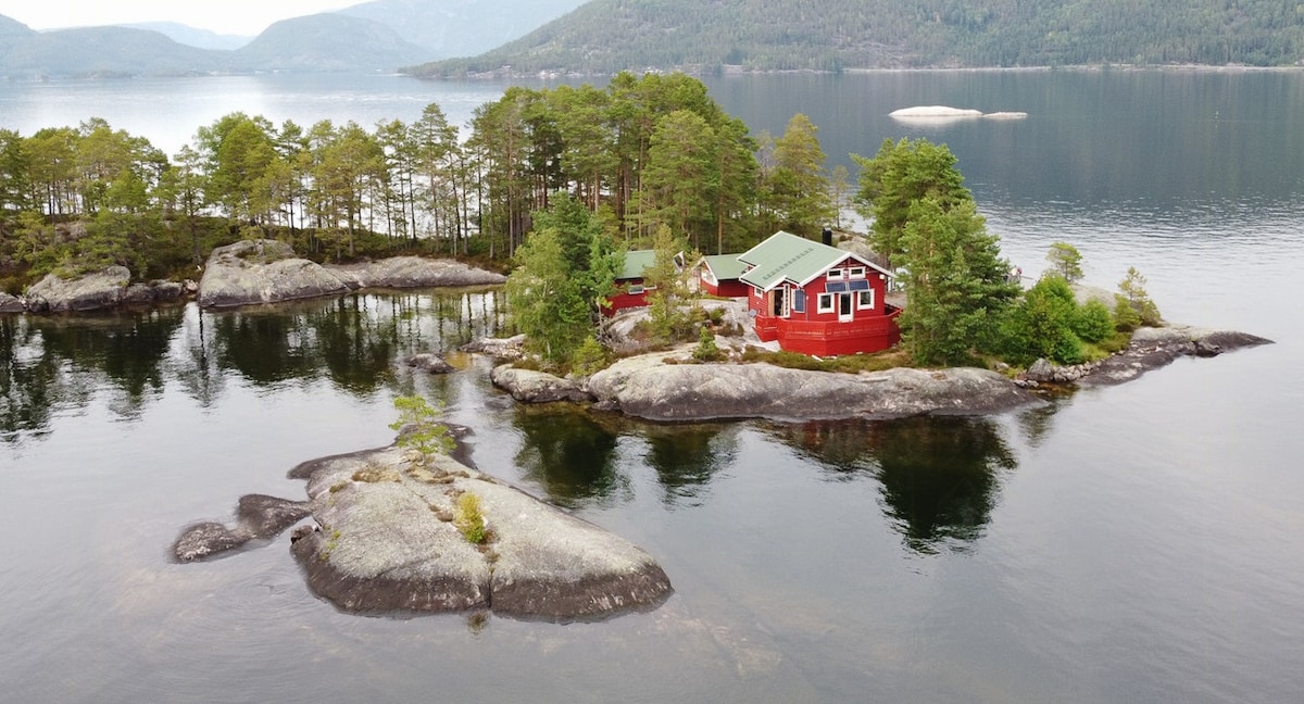A red cabin with a green roof is situated on a private island surrounded by calm water and rocky outcrops. Lush green trees dot the landscape, creating a serene backdrop against the distant mountains and open sky.