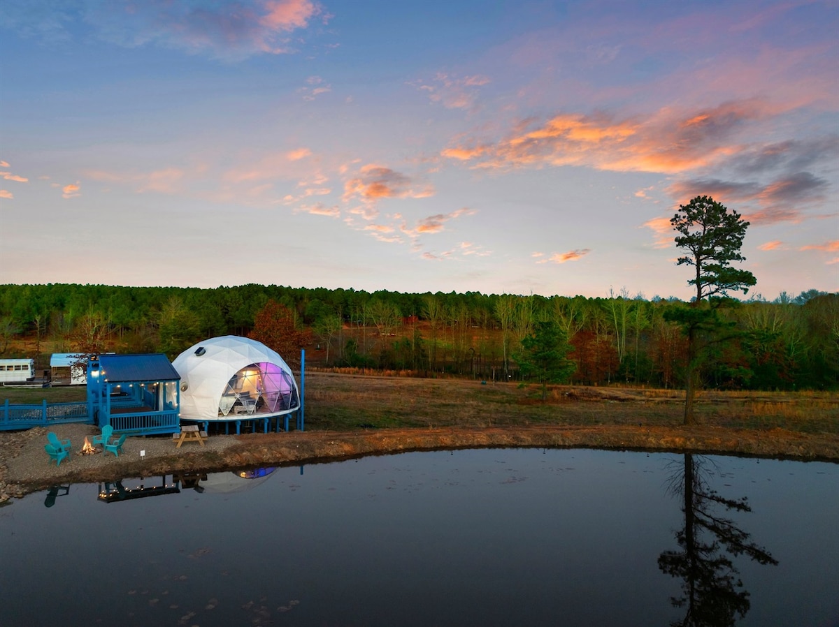The geodesic dome is positioned near a serene pond, with a blue deck leading to its entrance. Vibrant sunset hues paint the sky above a forested backdrop, while three blue chairs are arranged near the water's edge, inviting guests to relax and enjoy the peaceful surroundings.