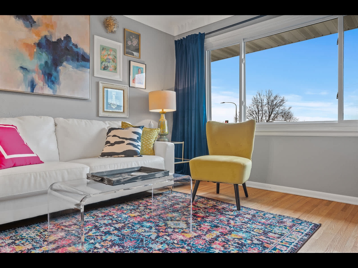 A light-filled living area features a white sofa adorned with colorful pillows and a decorative abstract artwork on the wall. A vibrant area rug adds character, while a yellow accent chair provides contrast. Large windows allow for ample natural light and views of the outdoors.