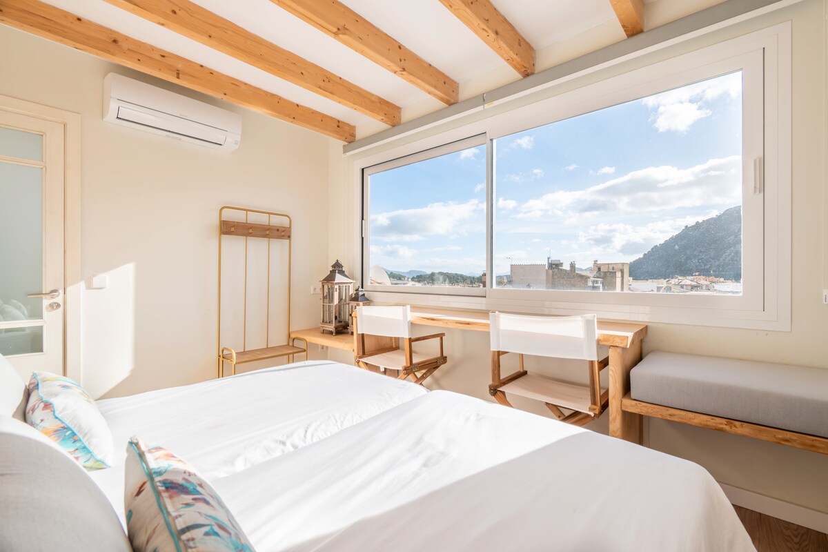 A bright bedroom features large windows presenting a view of the mountains and sky. Natural light fills the space, highlighting wooden ceiling beams and a cozy seating area. A workspace with a wooden desk and a chair is positioned along one wall, complementing the serene atmosphere.