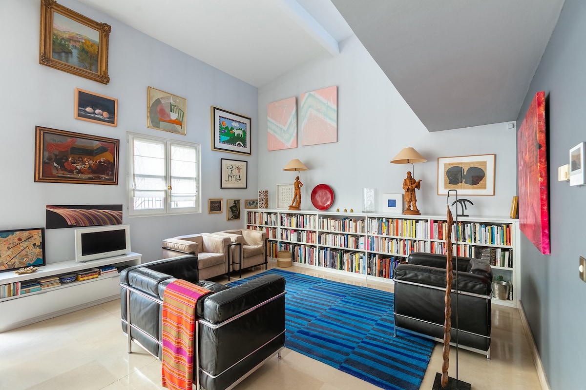 A spacious lounge area features wall art arranged in an eclectic display. A bookshelf filled with books occupies one wall, complemented by a television and a cozy armchair. Light from two windows brightens the room, while a striped rug adds a touch of color underfoot.