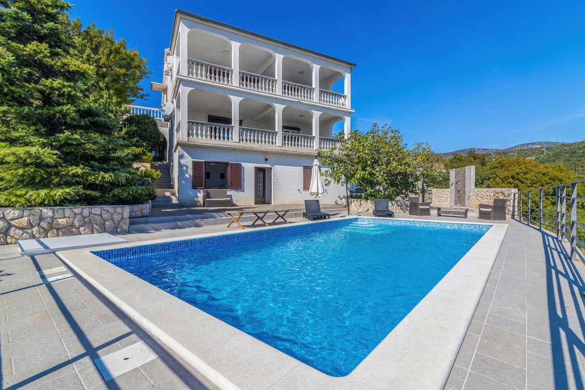 The image captures a three-story holiday home featuring multiple balconies and a well-maintained garden. A private swimming pool occupies the foreground, surrounded by lounge chairs, with clear blue water inviting relaxation. In the background, lush greenery and distant hills are visible under a clear blue sky.