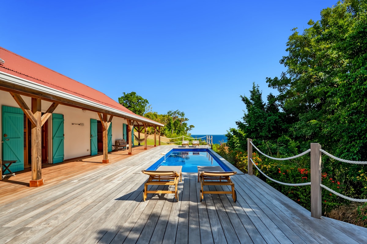 The exterior of the villa features a spacious wooden terrace lined with tropical greenery, leading to a swimming pool with a serene sea view. Comfortable lounge chairs are positioned poolside, inviting relaxation under clear blue skies.