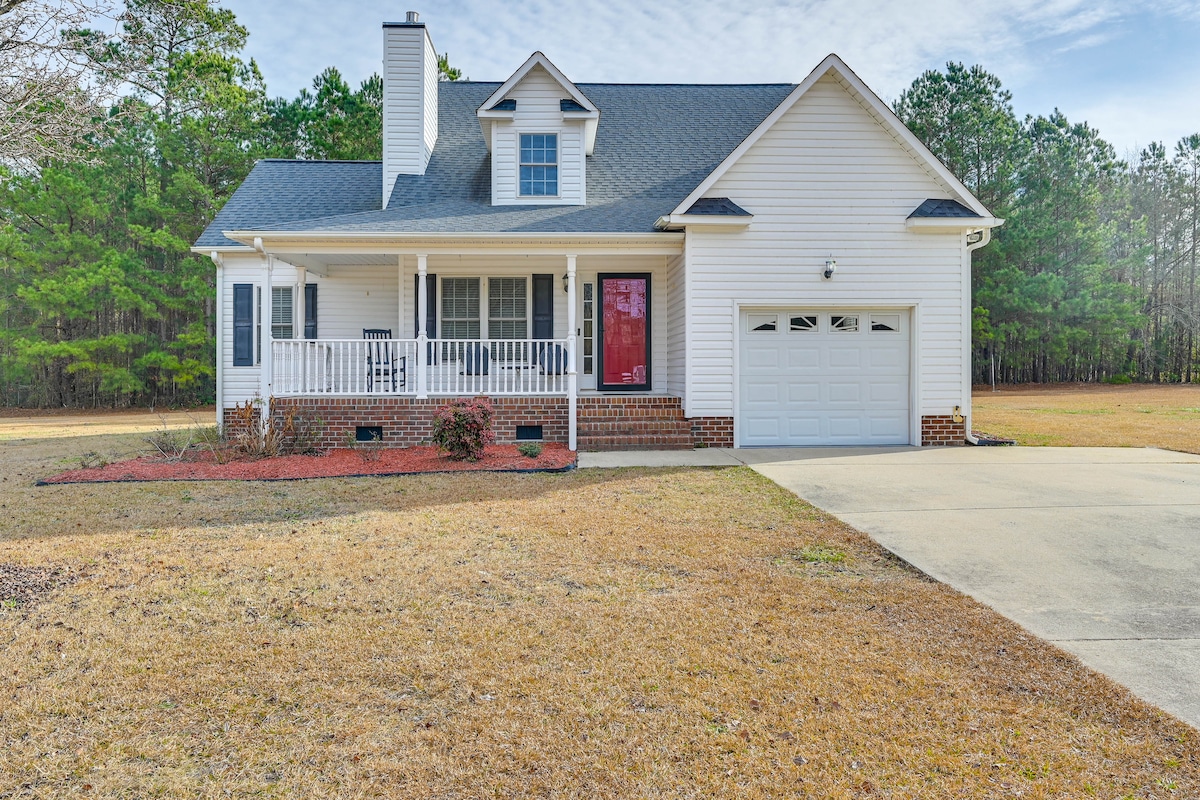 A charming two-story home is presented, featuring a welcoming porch with white railings and a red front door. A neatly paved driveway leads to a single-car garage, surrounded by landscaped areas. Lush green trees are visible in the background, providing a natural setting.