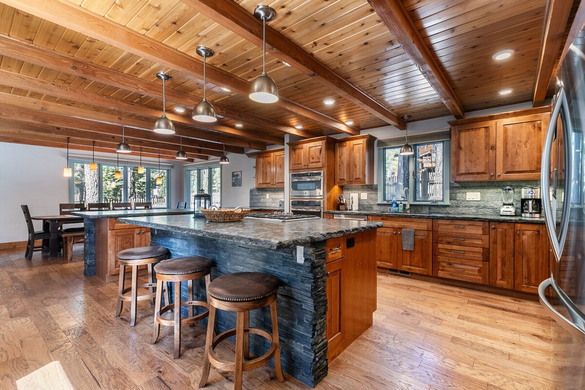 The kitchen features a large interactive island with granite countertops, surrounded by wooden stools. Rich wood cabinetry lines the walls, while stainless-steel appliances are integrated seamlessly. A warm wood ceiling adds to the inviting atmosphere, and large windows provide natural light and views of the outdoors.