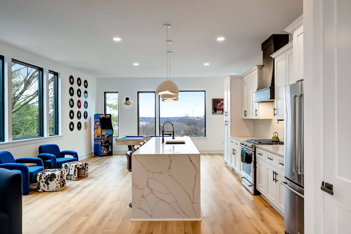 A modern kitchen is showcased with sleek cabinetry and a large kitchen island at its center. Sunlight fills the space through expansive windows, revealing inviting seating areas. A game table is positioned nearby, adding leisure to the culinary atmosphere.