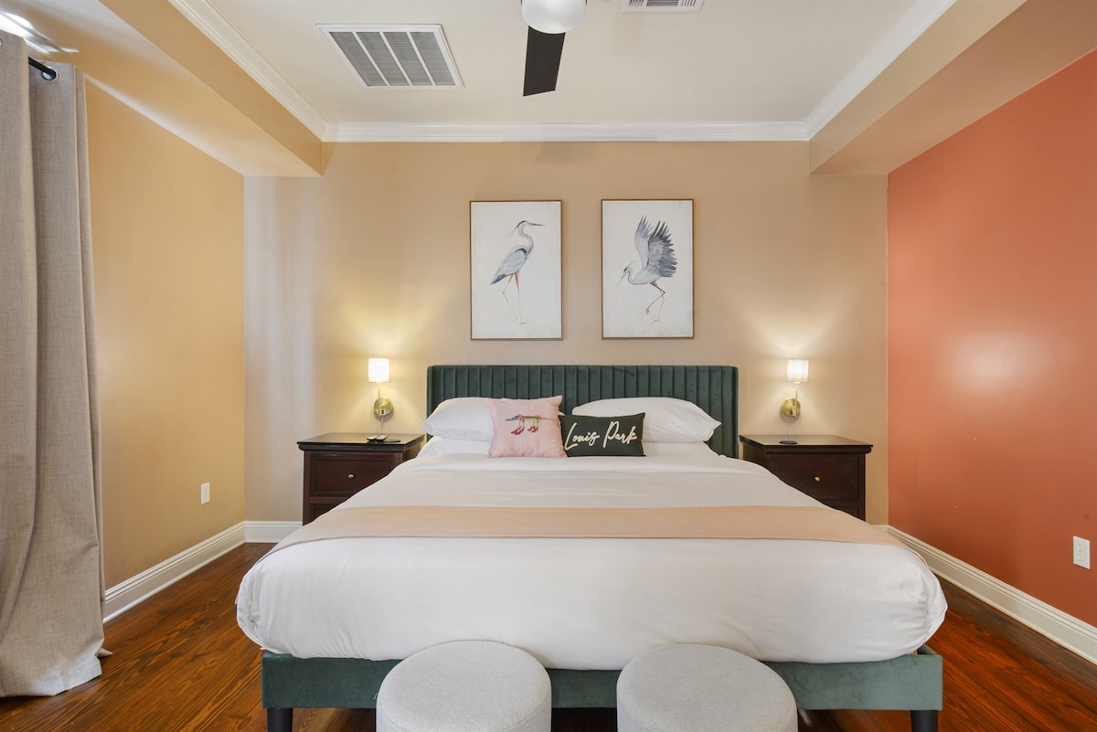A spacious bedroom showcases a king bed with premium linens. Two framed bird prints adorn the walls above the bed, while dual bedside lamps provide soft lighting. A low bench sits at the foot of the bed, and warm earth-toned walls contribute to the inviting ambiance.