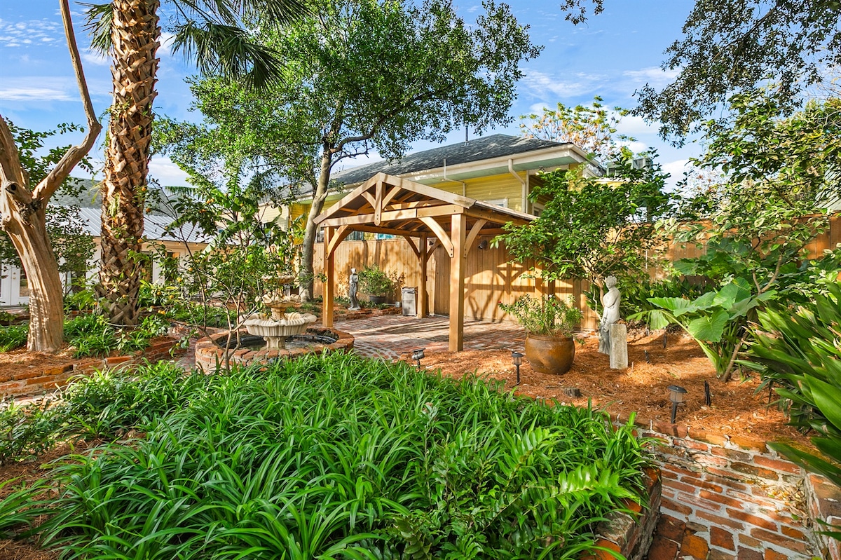 A landscaped courtyard showcases lush greenery and vibrant plants, framed by mature palm and shady trees. A wooden pergola offers a sheltered seating area, while soft paths made of brick wind through the garden, inviting leisurely strolls amongst the serene surroundings.