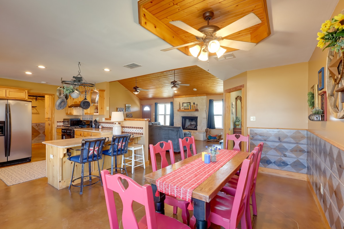 An open-plan living area showcases a well-equipped kitchen and a dining space. The wooden dining table is surrounded by vibrant red chairs. A ceiling fan and warm wood accents contribute to a welcoming atmosphere, while large windows allow natural light to fill the room.