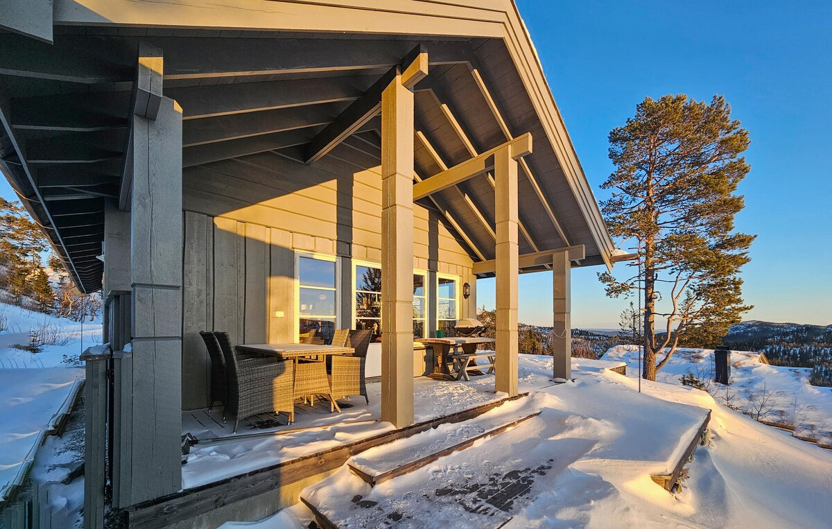 A spacious terrace features sturdy outdoor furniture, surrounded by a winter landscape covered in snow. Large windows allow for panoramic views, while a tree stands nearby, framing the inviting area. The structure's roof showcases a modern design with prominent beams.