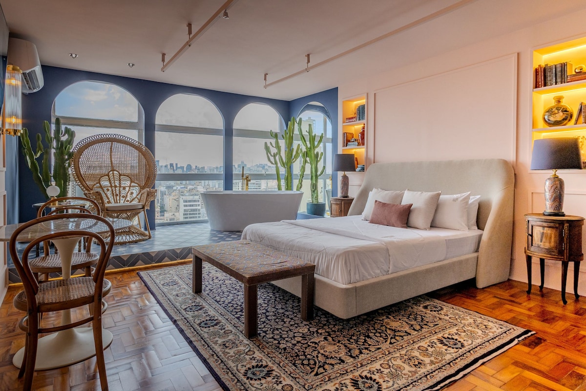 A stylish bedroom features a spacious bed with soft linens and a wooden table in the center. A cozy chair adds to the seating area, while large windows frame a scenic city view. A bathtub is positioned by the window, complementing the cactus decor.