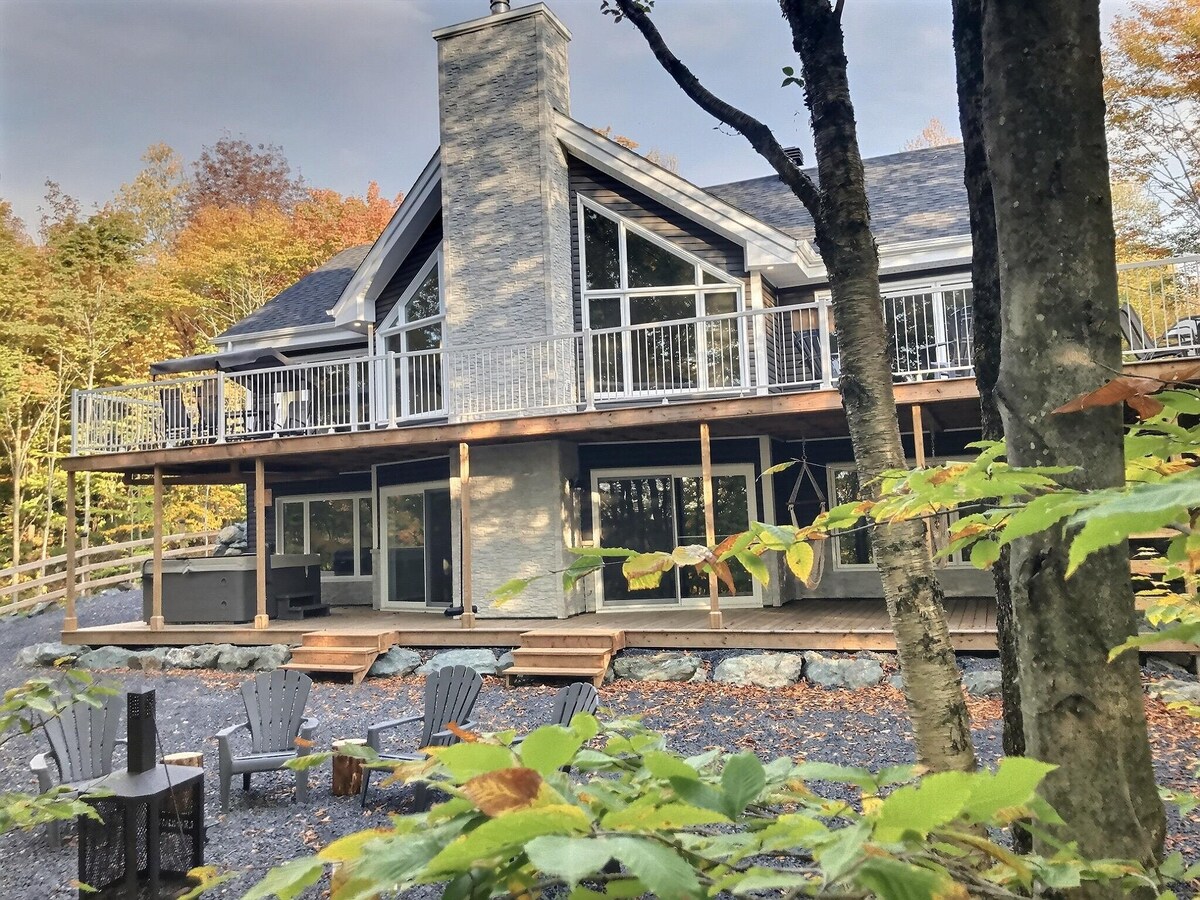 The chalet's exterior is showcased, highlighting a large deck with glass railings and a chimney. The surrounding trees display autumn foliage, enhancing the natural setting. A gravel area features several adirondack chairs and a fire pit, creating an inviting space for relaxation.