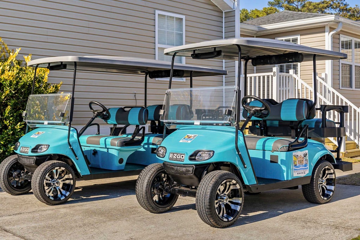 Two modern gas-powered golf carts are parked in front of the duplex, featuring a vibrant turquoise color with black and teal seats. The carts are equipped with sleek wheels and are ready for exploration, providing easy access to nearby attractions.