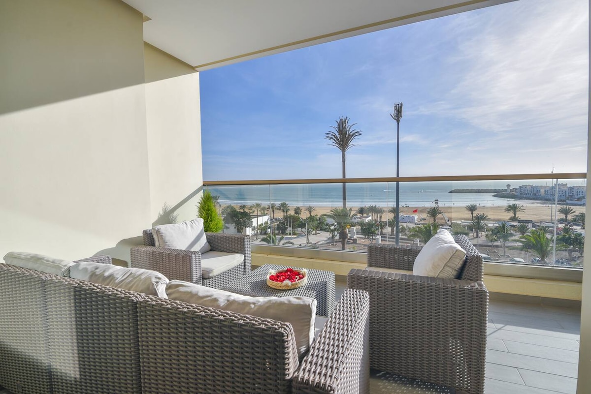 An inviting terrace features comfortable wicker seating, with a central table adorned with a decorative bowl. Panoramic views of the ocean and palm-lined coast are visible through expansive glass railings, providing a serene outdoor space for relaxation.