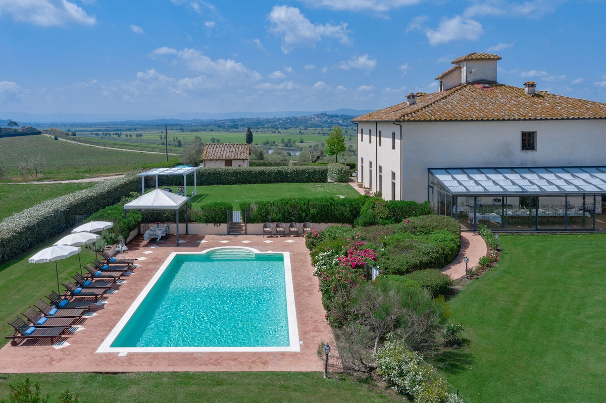 A scenic outdoor pool is framed by a lush lawn and flowering gardens, with sun loungers arranged around it. A gazebo provides shaded seating, while a glassed-in verandah is visible attached to the villa, offering views of the surrounding countryside.