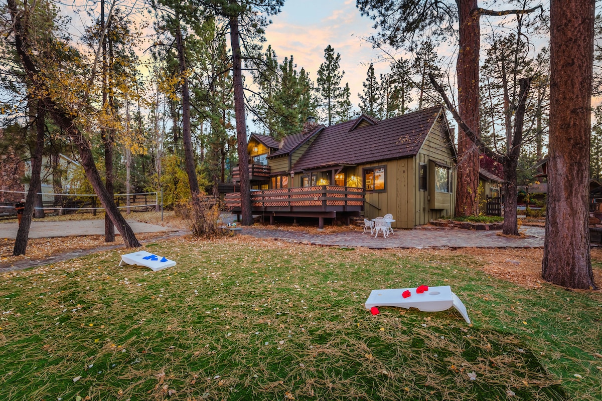 The exterior of a cabin is set among tall trees, with a spacious deck that features outdoor seating. Two cornhole boards are placed on the green lawn, while the rugged charm of the surroundings is complemented by a gentle glow from the evening sky.