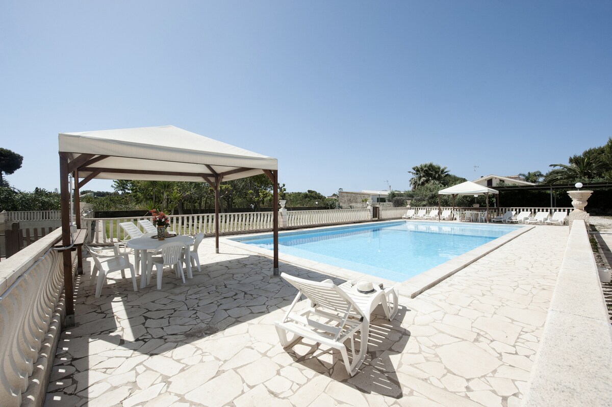A spacious swimming pool is surrounded by a well-maintained stone patio. Pairing sun loungers with a shaded dining area, the setting invites relaxation. Lush greenery borders the area, enhancing the tranquil environment.