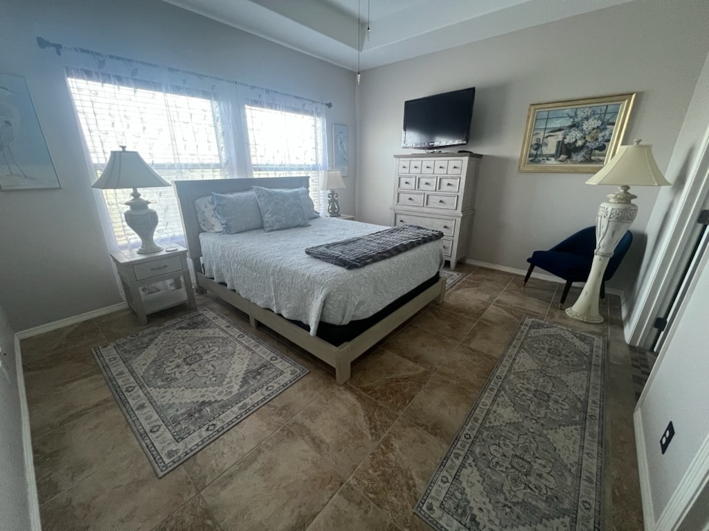 A well-appointed master bedroom features a spacious bed with a light-colored quilt. Two windows allow natural light, framed by sheer curtains. A sleek dresser and a mounted TV are present, accompanied by a small chair and elegant lamps on either side of the bed.