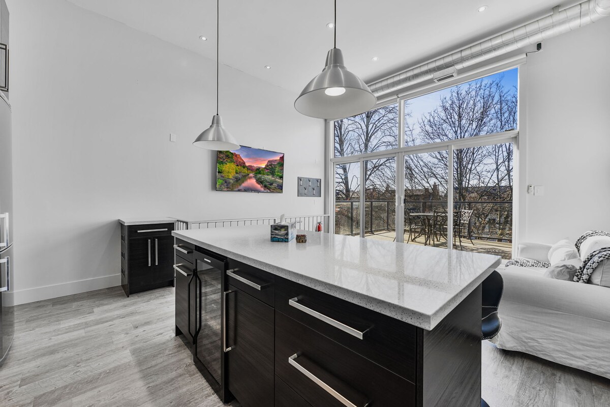 A modern kitchen features sleek, dark cabinetry and a spacious white countertop, illuminated by pendant lights. Floor-to-ceiling windows allow natural light to fill the area, offering views of the outdoor terrace. A cozy living space is present in the background, enhancing the open layout.