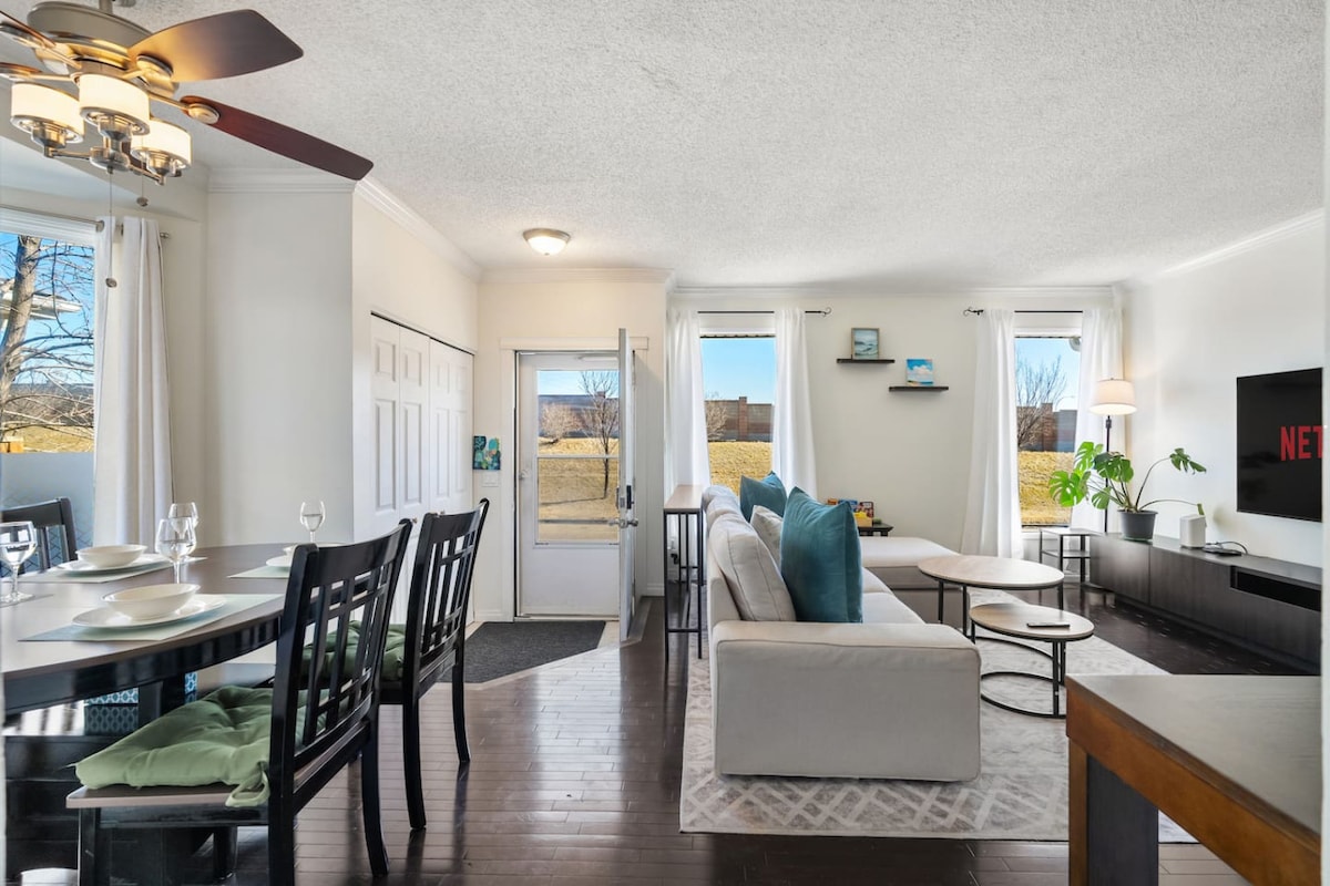 A bright living space is depicted, featuring a comfortable sofa adorned with blue cushions. A Smart TV is mounted on the wall, and dining arrangements for four are visible. Large windows invite natural light, enhancing the open atmosphere of the inviting area.