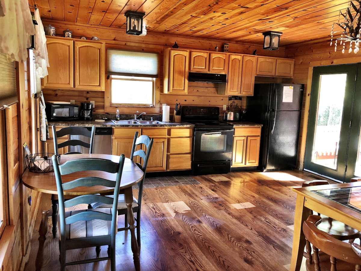 A warm wooden kitchen is highlighted by light-wood cabinetry and appliances in a dark finish. A round dining table with blue chairs is positioned near a window, providing natural light. Counter space is available with essential kitchen appliances and utensils visible.