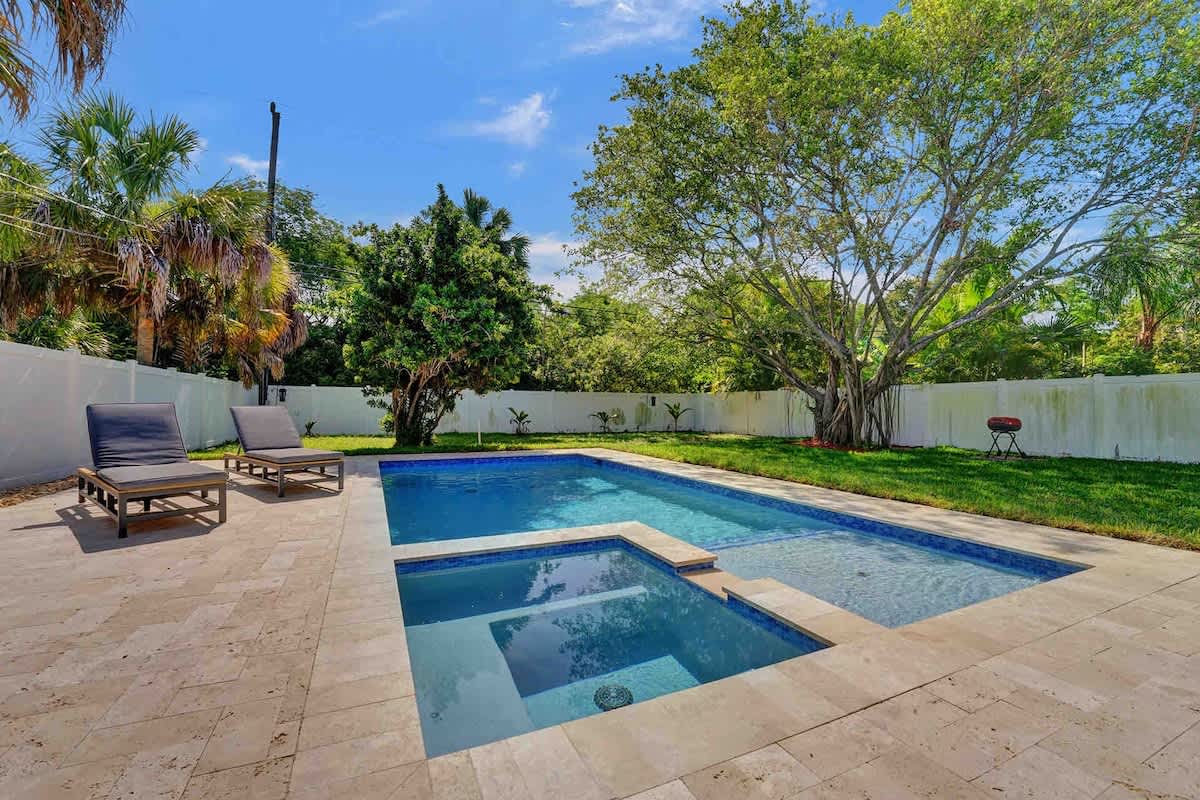 A serene outdoor area showcases a heated saltwater pool with a spa, surrounded by a well-maintained yard. Two lounge chairs are positioned nearby, inviting relaxation under the clear blue sky, while a lush tree adds natural shade to the setting.