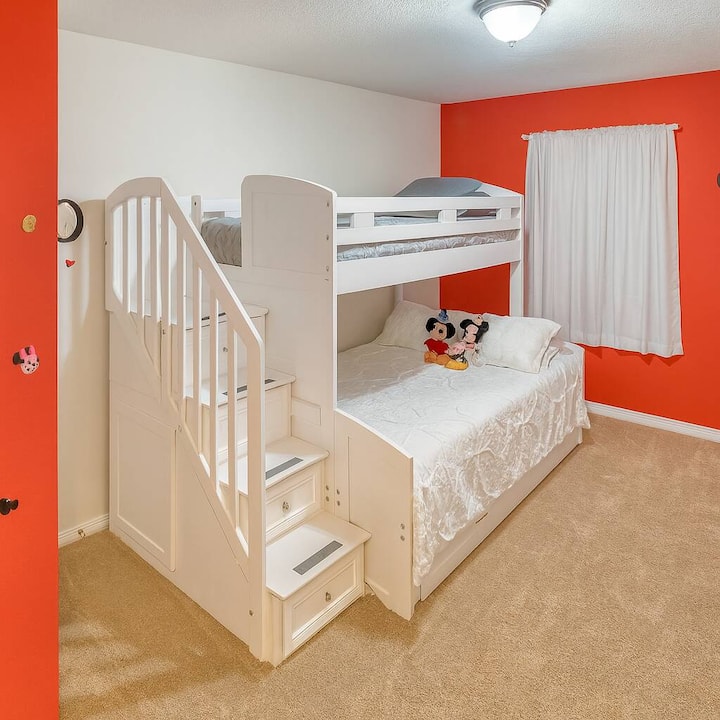 Deze gezellige kamer is licht en speels, met een charmant stapelbed met een gladde witte afwerking. De levendige oranje muur en zachte vloerbedekking creëren een vrolijke sfeer, versterkt door natuurlijk licht dat door het raam stroomt.