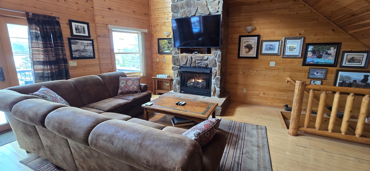 The open-concept living area features a comfortable sectional sofa in neutral tones, placed around a wooden coffee table. A stone gas fireplace is centered against a warm wooden wall, with a flat-screen television mounted above. Large windows allow natural light to illuminate the space.