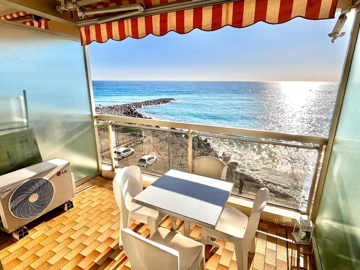 A terrace with a breathtaking sea view is presented, featuring a table and four chairs. The shimmering sea reflects sunlight, and an air conditioning unit is visible nearby. A serene atmosphere is created by the distant coastline and gentle waves lapping against the shore.