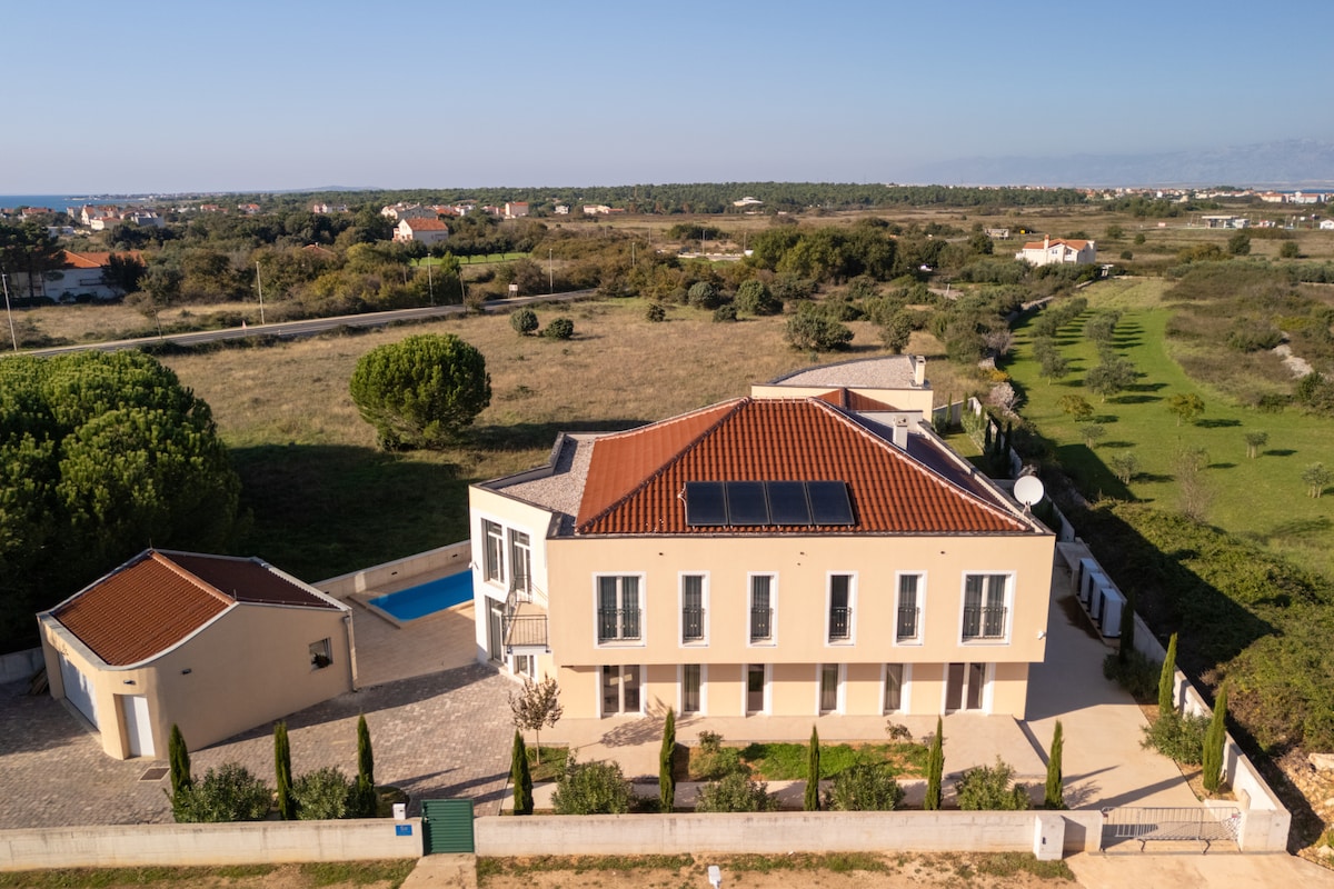 The exterior of Villa Ljubica is displayed, showcasing a spacious two-story building with a terracotta roof and modern design. An inviting swimming pool is visible to one side, surrounded by lush greenery and gentle landscaping. The surrounding area features open fields and a distant view of the coastline.