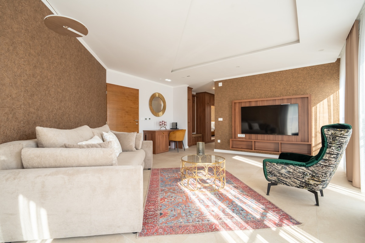 A modern sitting area is depicted with a plush sofa and an ornate chair. A decorative table is centered on an area rug, while a flat-screen TV is mounted on a wooden panel. Natural light streams in, highlighting the warm tones of the room.