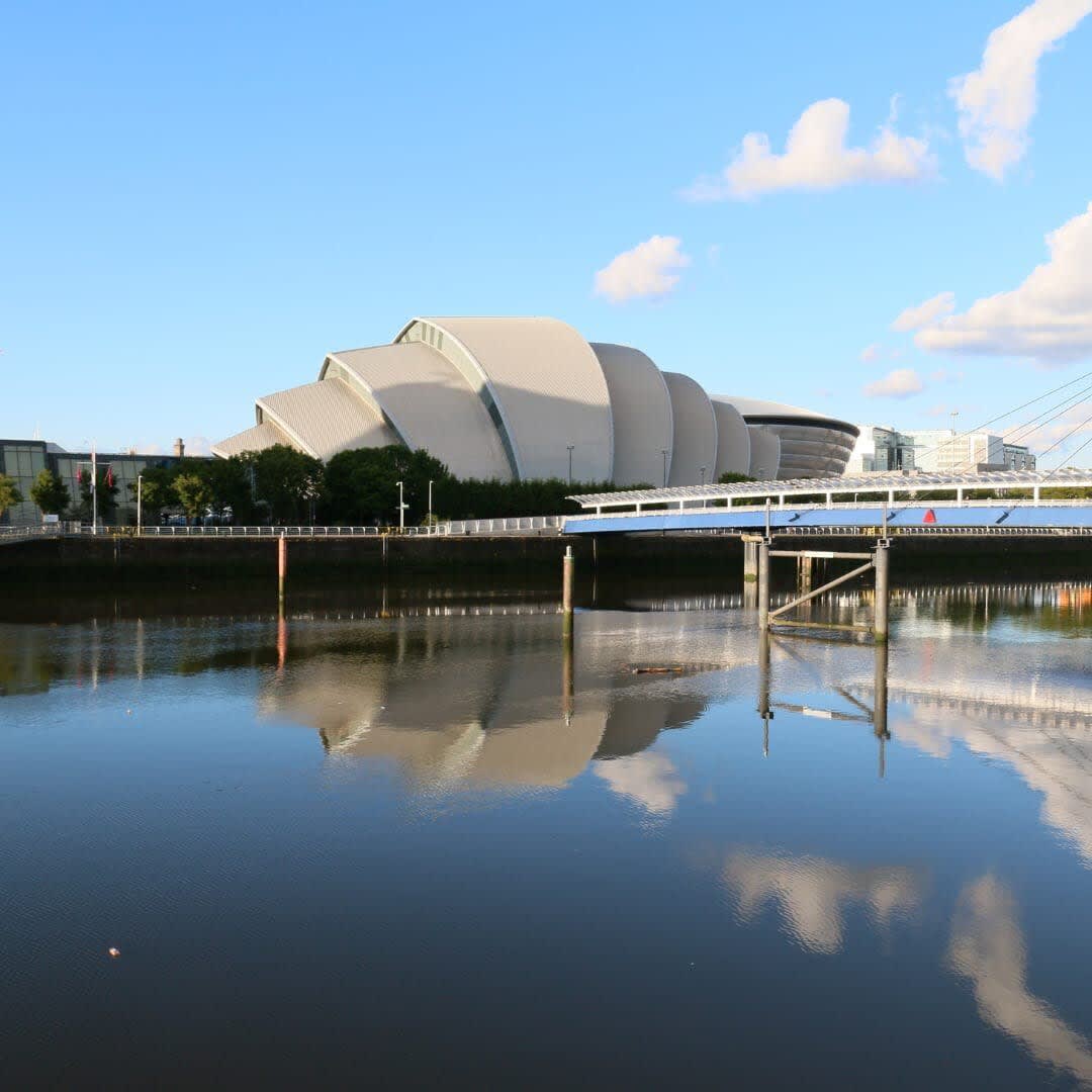 The image showcases the distinctive architecture of the Hydro events arena along the River Clyde. The building is reflected in the calm water, with a clear blue sky above and gentle clouds providing a serene backdrop.