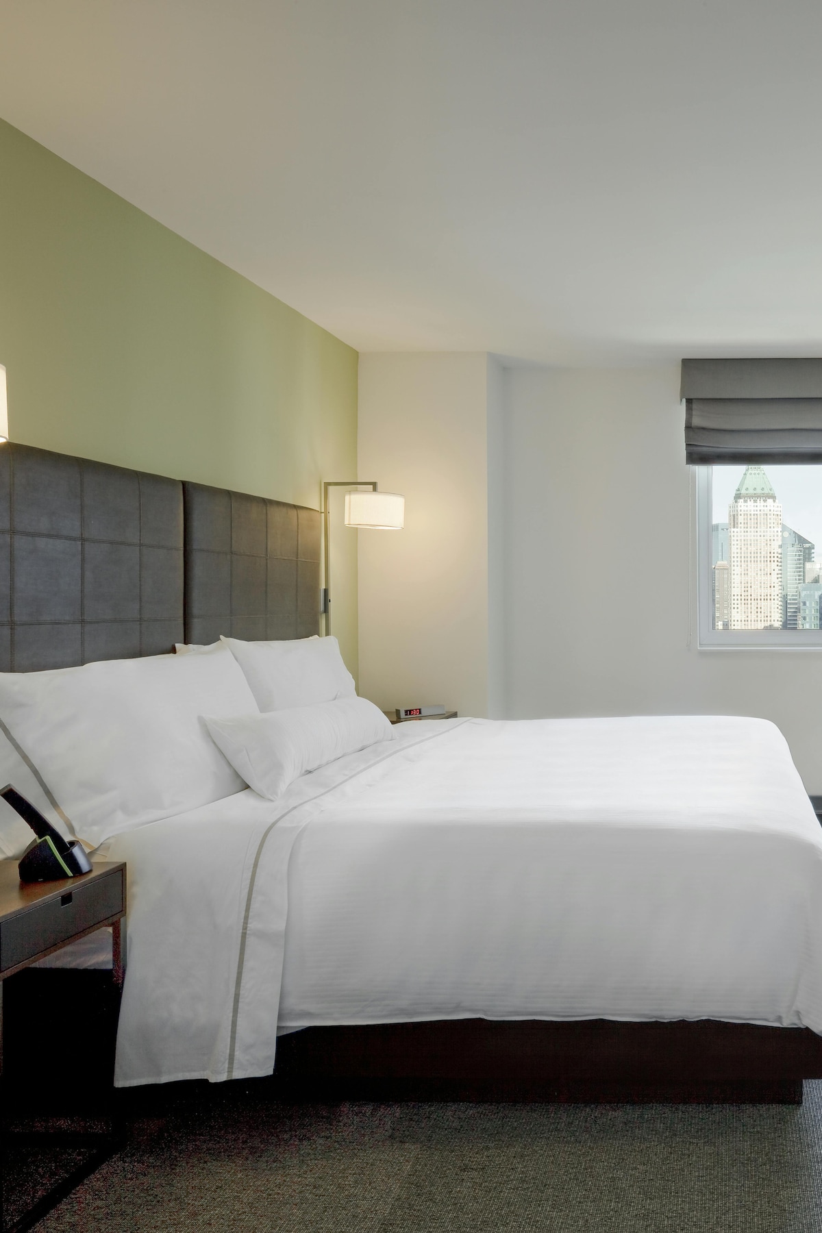 A modern room features a spacious bed with crisp white linens against a dark headboard. Light green walls create a calm atmosphere, while a large window provides a view of the city skyline. Functional bedside tables complement the contemporary design.