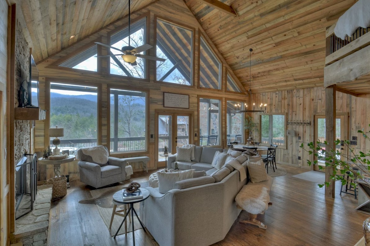 An open living area features vaulted wooden ceilings and large windows that allow natural light to illuminate the space. A comfortable sectional sofa and armchair are arranged around a coffee table, inviting relaxation while showcasing views of the mountains.
