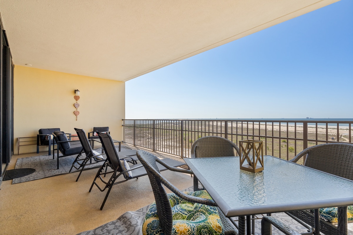 An expansive balcony features a dining table and several lounge chairs, inviting relaxation. The railing frames views of sandy beaches and ocean waves, while the floor is adorned with a patterned area rug. Sunlight highlights the outdoor furnishings against a clear blue sky.
