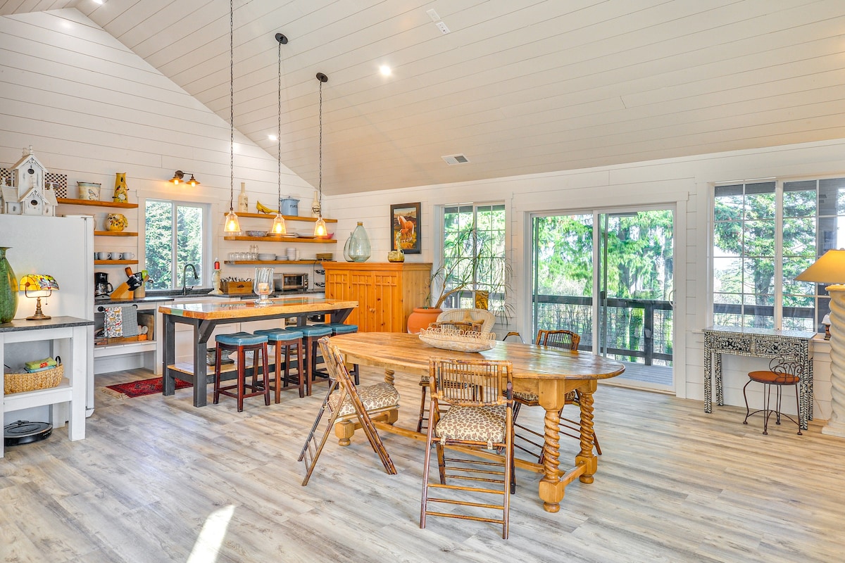 An open floor plan features a rustic dining area with a round wooden table surrounded by woven chairs. A kitchen with modern appliances, including a flat-screen TV and a cozy seating area, is visible. Large windows provide views of greenery, enhancing the bright, airy atmosphere.