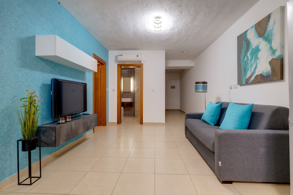 A modern living area features a sleek grey sofa adorned with turquoise cushions. A wall-mounted TV is positioned above a minimalist console table. The walls are painted a calming blue, complemented by a piece of abstract wall art. Natural light fills the space from an entrance opposite the seating area.