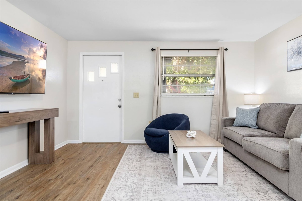 A welcoming living room features a gray sofa and a modern coffee table, complemented by a cozy blue chair. A large flat-screen TV is mounted on the wall, and natural light enters through a window adorned with light curtains. The space showcases wood-like flooring and neutral wall tones.