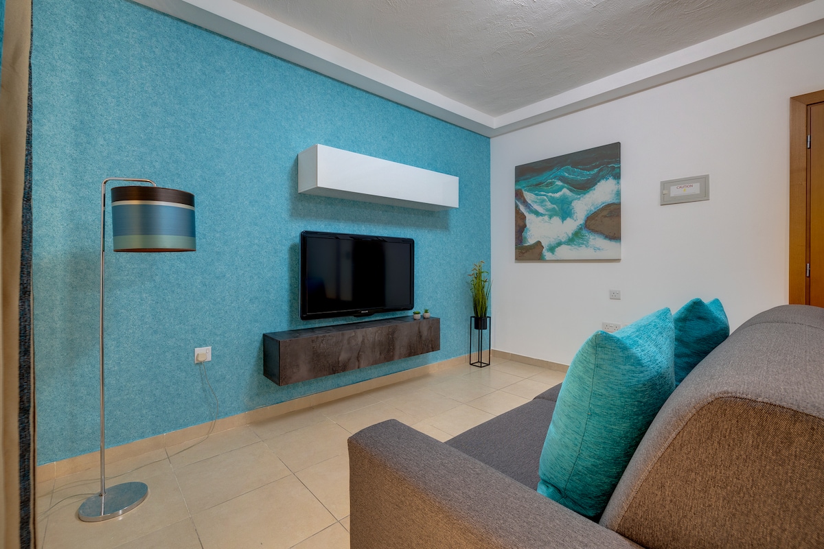 A modern living area features a grey sofa adorned with turquoise cushions, complemented by a floor lamp with a striped shade. A flat-screen TV is mounted on a minimalist wall shelf above a dark media console. A piece of abstract artwork adds a pop of color to the blue accent wall.