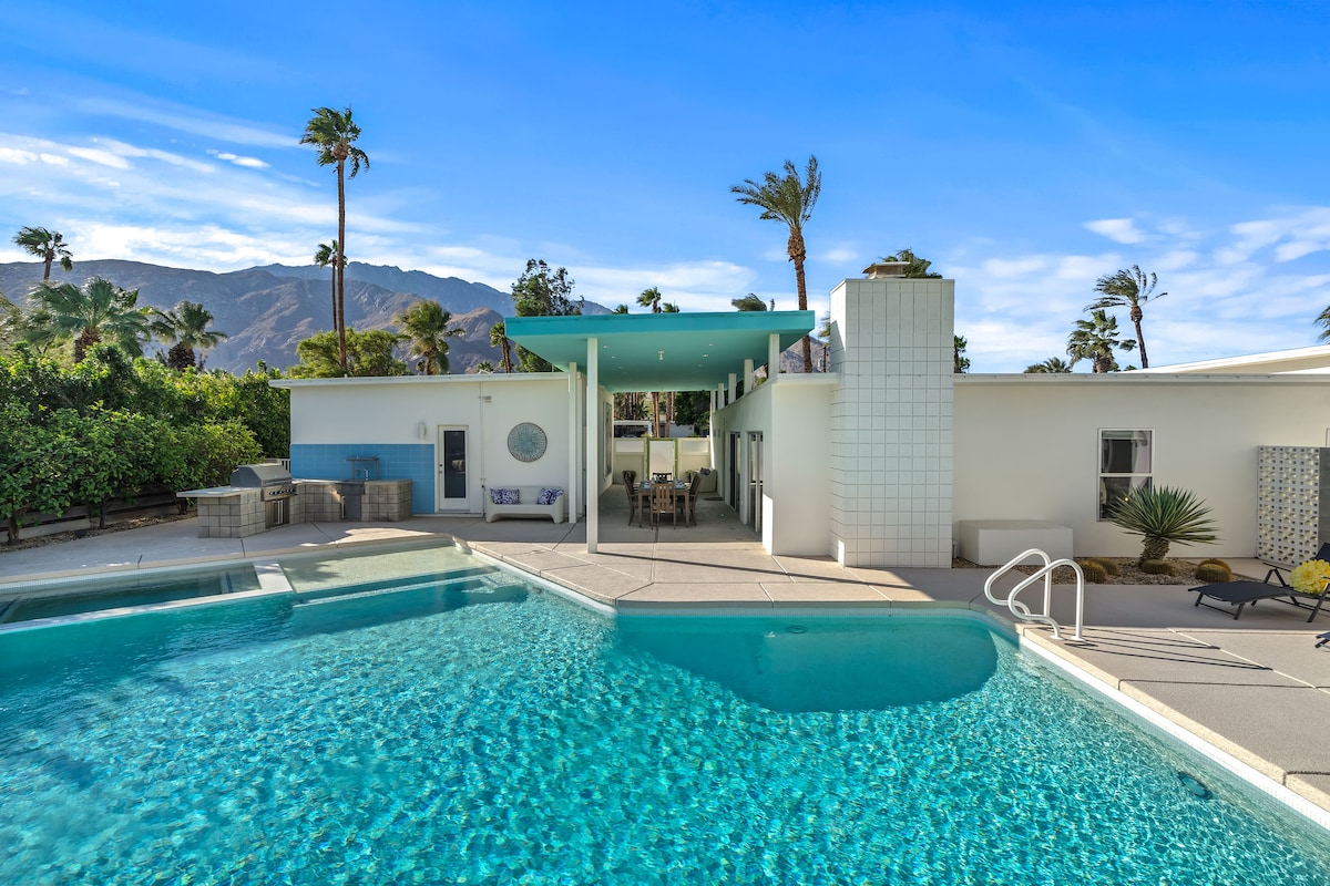 The image showcases a sparkling salt-water pool surrounded by a spacious deck. A stylish outdoor lounging area is visible under a shaded patio, with palm trees swaying gently in the background and mountains peeking through the lush vegetation.