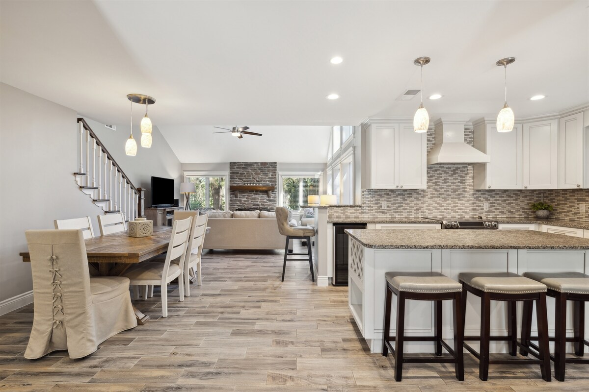 The open-concept living area features a modern kitchen with granite countertops and light cabinetry. A spacious dining table is set beside a cozy seating area that includes a flat panel TV. Abundant natural light floods the room, highlighting the elegant wood flooring.