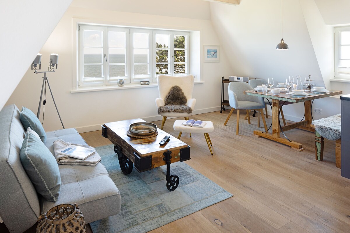 A bright and airy living space features a comfortable sofa with light blue cushions, a wooden coffee table on wheels, and a dining area with a wooden table set for four. Large windows provide a view of the sea, enhancing the room's openness.
