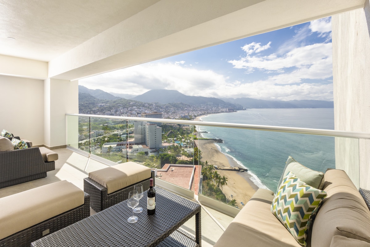 A spacious terrace is visible, featuring comfortable outdoor seating arrangements. A glass coffee table is positioned between two lounge chairs, with a bottle of wine and two glasses set atop it. The panoramic view showcases the coastline and mountains, framed by a clear sky.