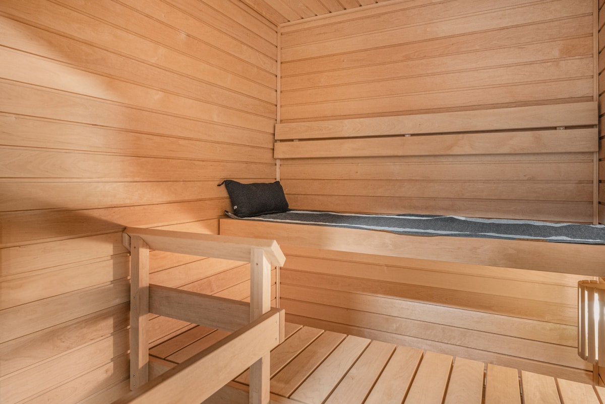 A minimalist sauna room is seen, featuring smooth wooden walls and benches. A narrow mattress is positioned on one of the benches, accompanied by a small cushion. Natural light filters through the space, providing a warm ambiance.