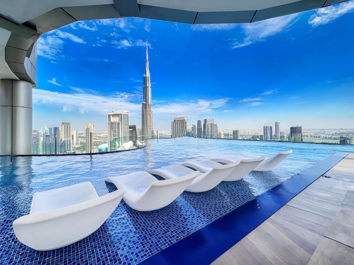 An adult pool area features sleek white loungers positioned along the edge of an infinity pool. The skyline of Dubai, including the Burj Khalifa, is visible in the background under a clear blue sky.