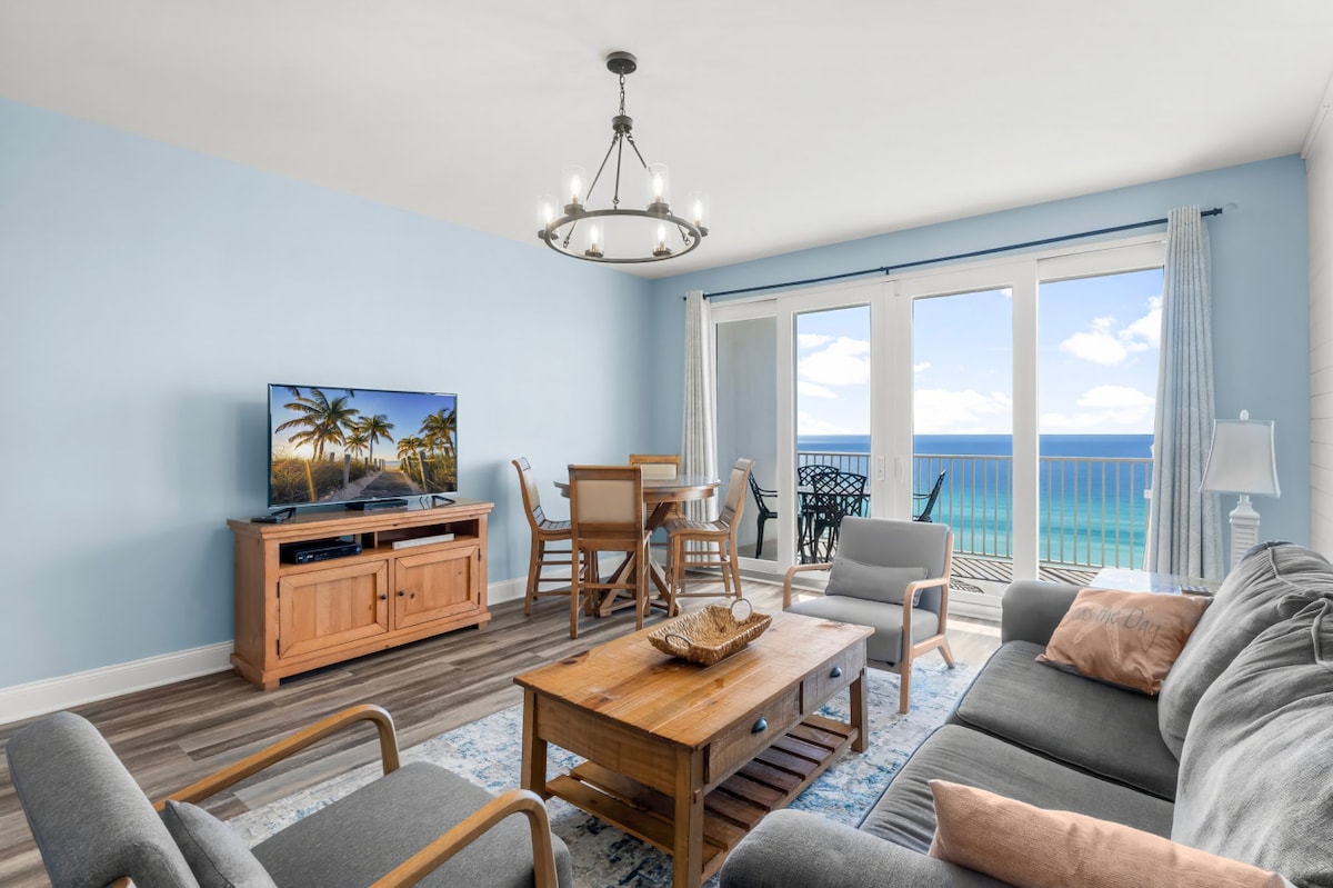 The living area features a comfortable sectional sofa, stylish armchairs, and a wooden coffee table. A TV is positioned on a media console, and large glass doors provide access to the balcony, offering panoramic ocean views. A dining table with chairs completes the space.