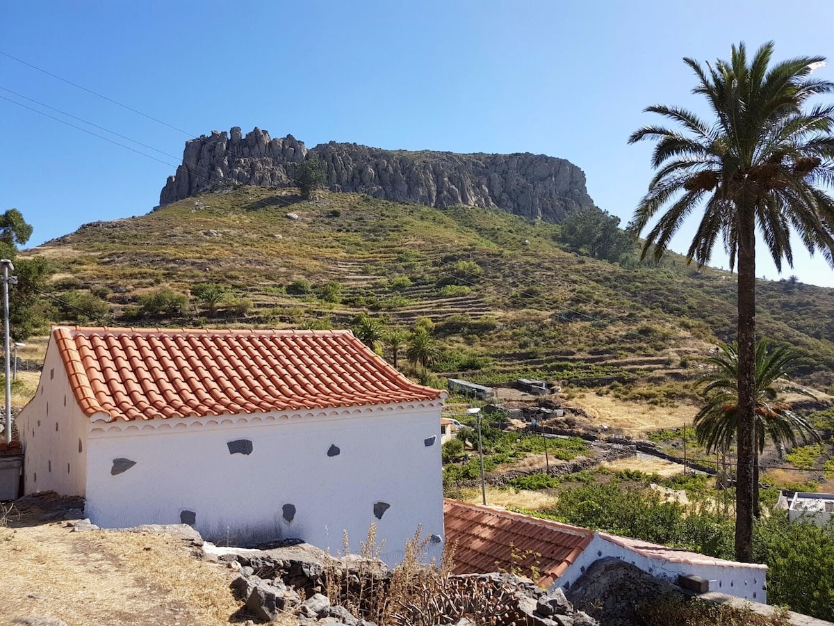 A white-walled house with a red-tiled roof is positioned in the foreground, framed by palm trees. In the background, the imposing La Fortaleza mountain rises under a clear blue sky, showcasing natural greenery and terraced land.